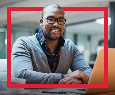 A Black developer wearing glasses smiles and sits in front of a laptop